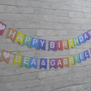 Puede incluir: Una pancarta de colores arco&iacute;ris con letras blancas que dice "Happy Birthday Bea & Gabriel". La pancarta tiene corazones blancos en cada extremo.