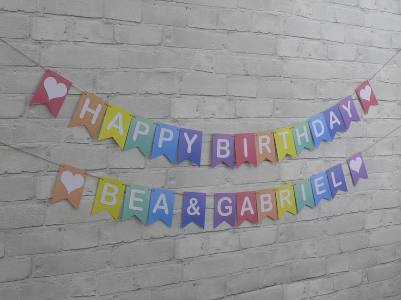 Puede incluir: Una pancarta de colores arco&iacute;ris con letras blancas que dice "Happy Birthday Bea & Gabriel". La pancarta tiene corazones blancos en cada extremo.