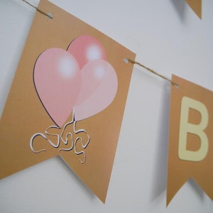 Op de afbeelding: Een bruine banner met het woord "Happy" en het woord "Birthday" in gele letters. De banner is versierd met roze ballonnen.
