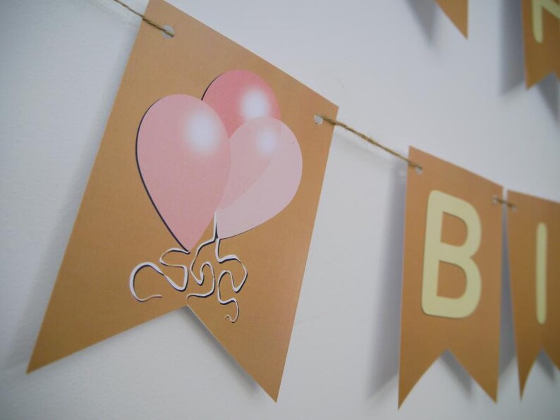 Op de afbeelding: Een bruine banner met het woord "Happy" en het woord "Birthday" in gele letters. De banner is versierd met roze ballonnen.