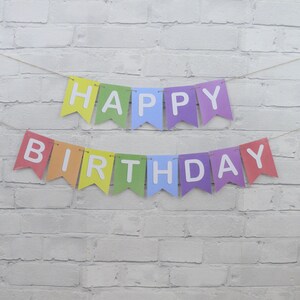 May include: A colorful "Happy Birthday" banner with rainbow-colored pennants and white lettering. The banner is strung on a thin twine against a white brick wall background. Perfect for birthday celebrations.