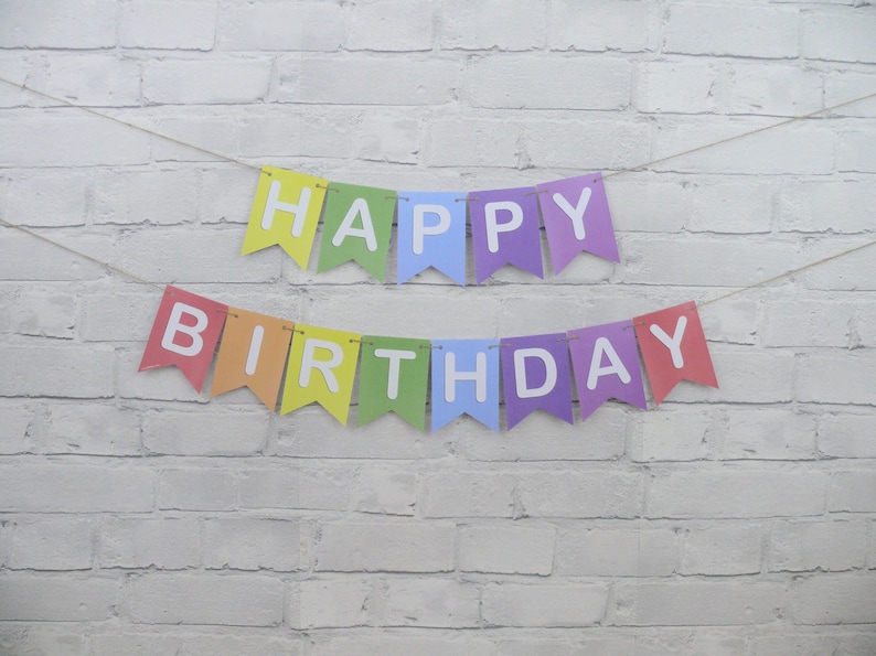 May include: A colorful "Happy Birthday" banner with rainbow-colored pennants and white lettering. The banner is strung on a thin twine against a white brick wall background. Perfect for birthday celebrations.