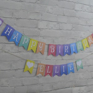 Puede incluir: Una pancarta de colores arco&iacute;ris con letras blancas que dice "Happy Birthday Ellie". La pancarta tiene corazones blancos entre cada letra.