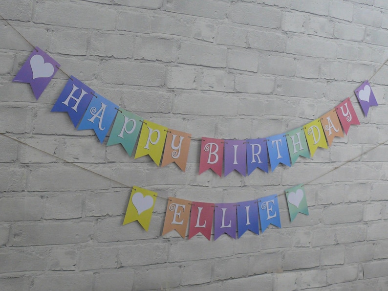 Puede incluir: Una pancarta de colores arco&iacute;ris con letras blancas que dice "Happy Birthday Ellie". La pancarta tiene corazones blancos entre cada letra.