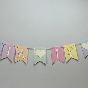 May include: Pastel colored banner with the text "Olivia is one" in white letters with a heart between "is" and "one".