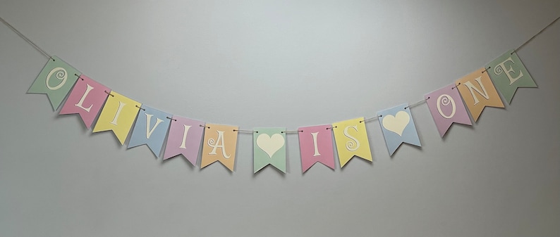 May include: Pastel colored banner with the text "Olivia is one" in white letters with a heart between "is" and "one".