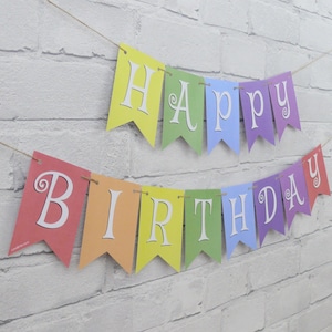 May include: A colorful banner with the words "HAPPY BIRTHDAY" strung across a white brick wall. Each pennant is a different color of the rainbow, with white letters. The banner is strung on a thin, natural-colored twine.