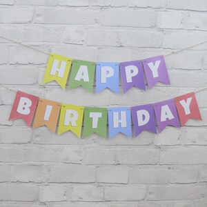 May include: A colorful "Happy Birthday" banner with individual pennants in rainbow hues. Each pennant displays a white letter, spelling out the message. The banner is strung on a thin, natural-colored twine, set against a white brick wall.