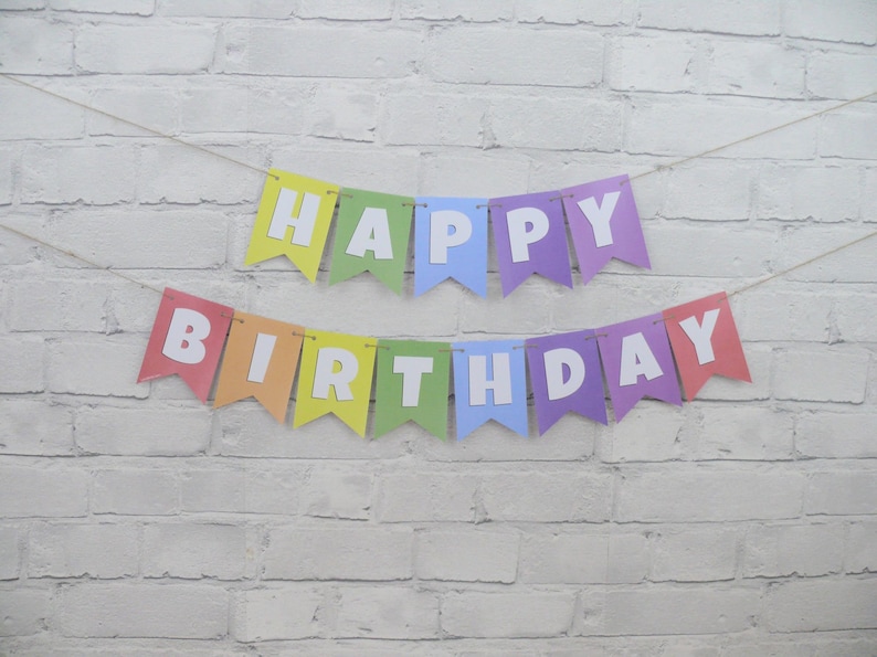 May include: A colorful "Happy Birthday" banner with individual pennants in rainbow hues. Each pennant displays a white letter, spelling out the message. The banner is strung on a thin, natural-colored twine, set against a white brick wall.