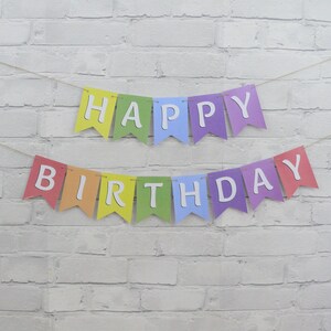 May include: A colorful "Happy Birthday" banner with individual pennants in rainbow colors. Each pennant displays a white letter, spelling out "HAPPY" on the top row and "BIRTHDAY" on the bottom row. The banner is strung on a thin, natural-colored twine.