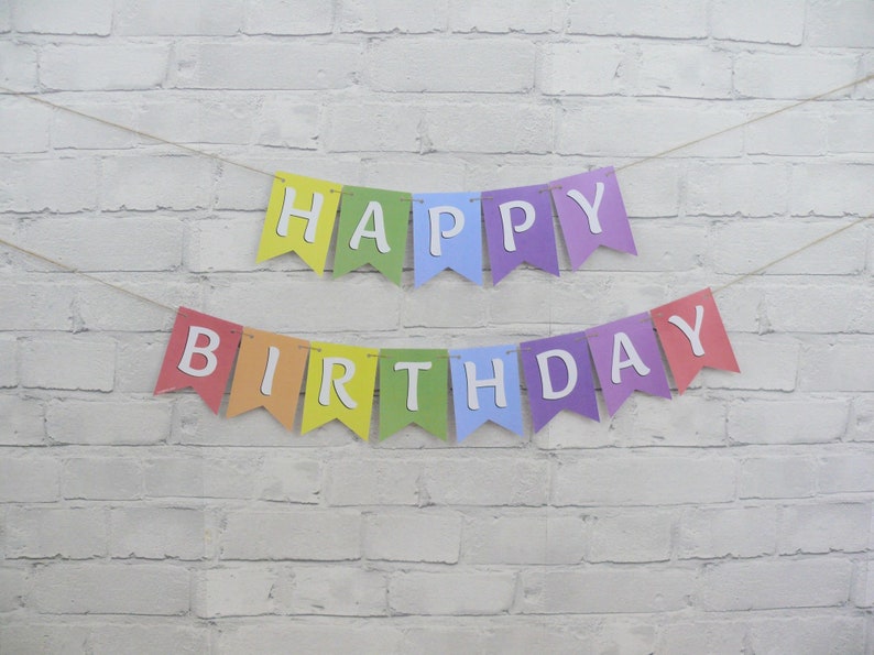 May include: A colorful "Happy Birthday" banner with individual pennants in rainbow colors. Each pennant displays a white letter, spelling out "HAPPY" on the top row and "BIRTHDAY" on the bottom row. The banner is strung on a thin, natural-colored twine.