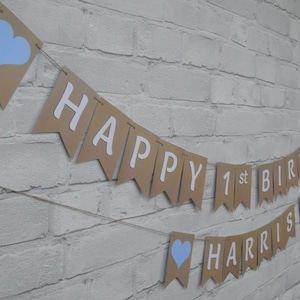 Op de afbeelding: Een verjaardagsbanner met bruine vlaggetjes met de tekst "HAPPY 1st BIRTHDAY HARRIS" in witte letters. Sommige vlaggetjes hebben lichtblauwe harten. De banner is geregen aan een touw en wordt weergegeven tegen een witte bakstenen muur.