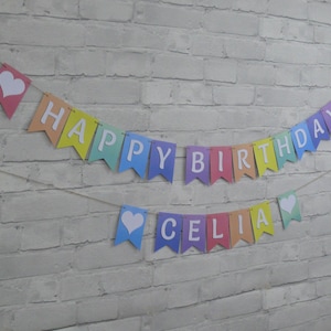 Puede incluir: Una pancarta de colores arco&iacute;ris con letras blancas que dice "Happy Birthday Celia" con corazones blancos en cada extremo.
