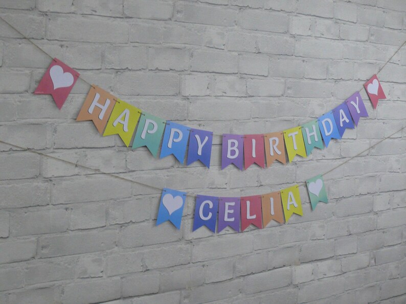 Puede incluir: Una pancarta de colores arco&iacute;ris con letras blancas que dice "Happy Birthday Celia" con corazones blancos en cada extremo.