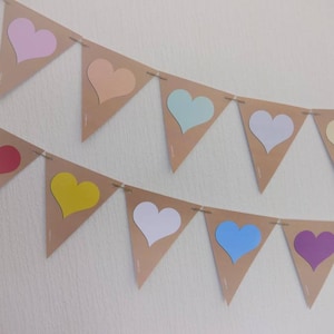 May include: Two rows of heart-shaped bunting. Each pennant is a light brown color with a heart cut out, revealing a pastel heart in pink, peach, mint, white, and yellow. The second row has a red, yellow, white, blue, and purple heart.