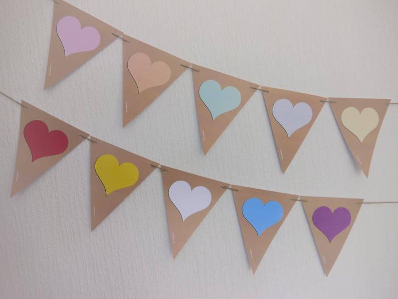 May include: Two rows of heart-shaped bunting. Each pennant is a light brown color with a heart cut out, revealing a pastel heart in pink, peach, mint, white, and yellow. The second row has a red, yellow, white, blue, and purple heart.