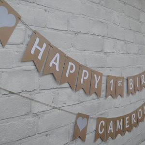 Op de afbeelding: Een bruine slinger met witte letters met de tekst "HAPPY 1st BIRTHAY CAMERON". De vlaggetjes zijn geregen aan touw en hebben witte harten. De slinger hangt tegen een witte bakstenen muur.