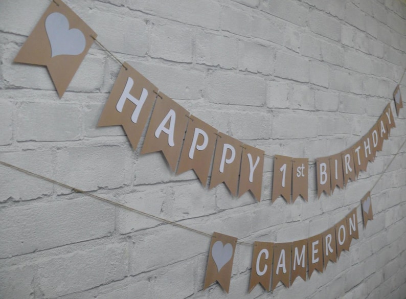 Op de afbeelding: Een bruine slinger met witte letters met de tekst "HAPPY 1st BIRTHAY CAMERON". De vlaggetjes zijn geregen aan touw en hebben witte harten. De slinger hangt tegen een witte bakstenen muur.