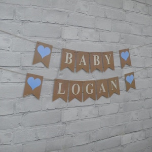 May include: A banner with the words "BABY LOGAN" strung across a twine. The banner is made of brown paper pennants with blue heart accents. The background is a white brick wall.