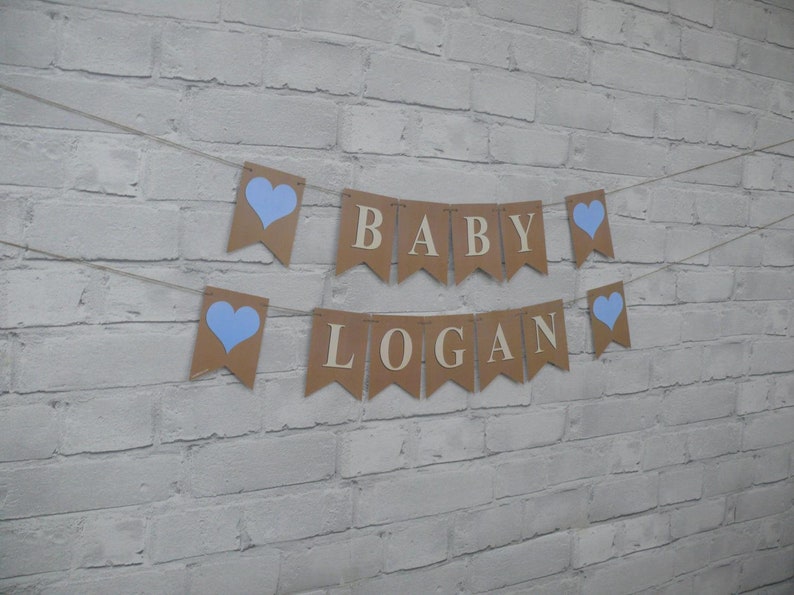 May include: A banner with the words "BABY LOGAN" strung across a twine. The banner is made of brown paper pennants with blue heart accents. The background is a white brick wall.