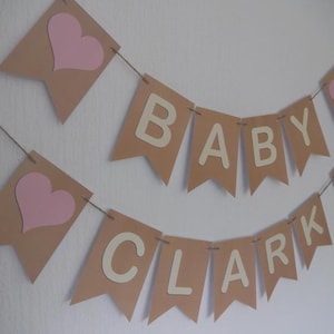 May include: A banner set for a baby shower, featuring pennants in tan with pink hearts and the words "BABY CLARK" in cream-colored letters. The banner is strung on twine and is against a white background.