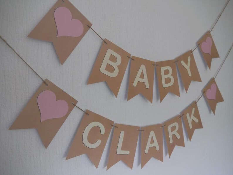 May include: A banner set for a baby shower, featuring pennants in tan with pink hearts and the words "BABY CLARK" in cream-colored letters. The banner is strung on twine and is against a white background.