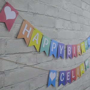 Puede incluir: Una pancarta de colores arco&iacute;ris con letras blancas que deletrean "Feliz cumplea&ntilde;os" y una pancarta m&aacute;s peque&ntilde;a con letras blancas que deletrean "Celia". Ambas pancartas tienen corazones blancos en cada extremo.