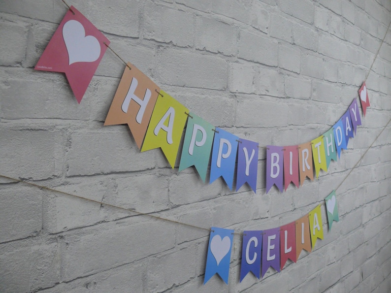 Puede incluir: Una pancarta de colores arco&iacute;ris con letras blancas que deletrean "Feliz cumplea&ntilde;os" y una pancarta m&aacute;s peque&ntilde;a con letras blancas que deletrean "Celia". Ambas pancartas tienen corazones blancos en cada extremo.