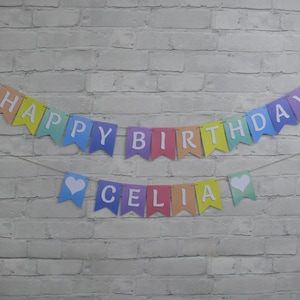 Puede incluir: Bandera arco&iacute;ris con letras blancas que deletrean "Happy Birthday Celia" con corazones blancos en cada extremo.