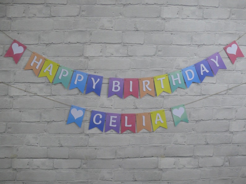 Puede incluir: Bandera arco&iacute;ris con letras blancas que deletrean "Happy Birthday Celia" con corazones blancos en cada extremo.