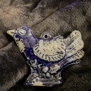 May include: A ceramic bird figurine in shades of blue and gray. The bird has a detailed design with a textured surface and a hole for hanging. The figurine is set against a dark, textured background.