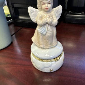 May include: A ceramic angel figurine with outstretched wings, holding a small object, atop a cloud-shaped trinket box. The box is white with gold trim and star accents. The angel has a light skin tone and is wearing a light-colored dress.