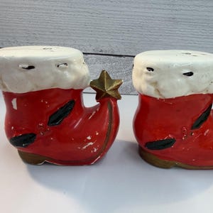May include: A pair of ceramic salt and pepper shakers shaped like red Christmas boots with white trim and gold star accents. The boots have black details and a distressed finish.