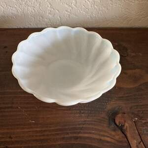 May include: A white, scalloped-edge bowl with a swirling design. The bowl is made of a translucent material, and sits on a dark wooden surface. The bowl's edges have a subtle blue tint.
