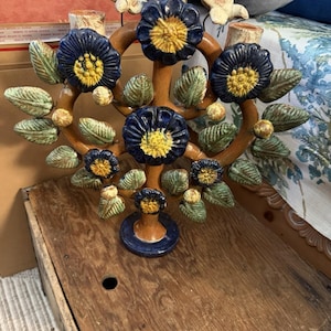 May include: A ceramic candelabra featuring a brown, tree-like structure adorned with dark blue flowers with yellow centers, green leaves, and small round buds. Two white bird figures are perched at the top near the candle holders.