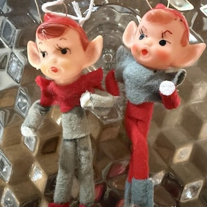 May include: Two vintage Christmas elf ornaments. Each elf has a painted face with rosy cheeks, red hair, and large ears. They wear red and gray felt outfits with matching hats and boots. The ornaments are suspended by thin wire and white string.