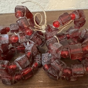 May include: Two decorative napkin rings made of dark red, square-cut glass beads and smaller red round beads. The beads are strung together with a natural twine. The napkin rings are on a wooden surface.