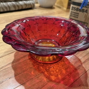 May include: A vibrant red glass bowl with a scalloped rim and a textured interior. The bowl sits on a golden-colored base, casting a warm shadow on the wooden surface. The bowl's design features a diamond pattern.