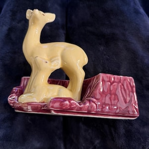 May include: A ceramic figurine of a deer in a light yellow color, standing on a burgundy base. The deer is looking upwards. The base has a rectangular shape with a textured surface.