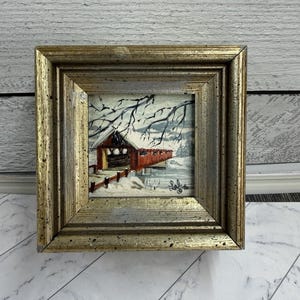 May include: A framed painting of a red covered bridge in a winter landscape. The artwork features a snow-covered scene with bare tree branches and a wooden fence. The frame is gold-colored.