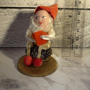 May include: A vintage figurine of a gnome with a red hat and shoes, holding a red book. The gnome has a white beard and is seated on a pinecone base atop a gold glittery disc. A ruler is next to the gnome.
