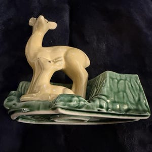 May include: A ceramic figurine of a deer in a cream color, with a green base and a tree stump. The deer is looking up. The base has a leaf design. The figurine is on a dark blue surface.