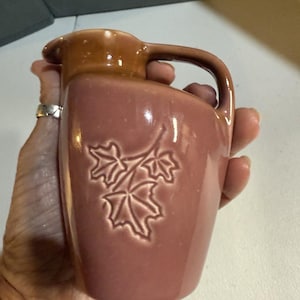 May include: A small, vintage pitcher in a muted rose color with a darker brown handle and spout. The pitcher features an embossed design of three maple leaves. The handle is curved, and the pitcher has a smooth, glossy finish.