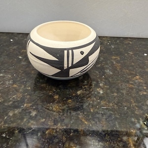 May include: A small, round ceramic bowl with a cream-colored interior and a black and white geometric design on the exterior. The bowl's design features angular shapes and lines. The bowl is sitting on a dark, speckled surface.