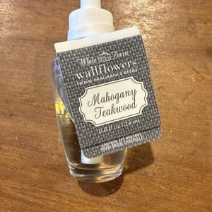 May include: A clear glass wallflower home fragrance refill with a white top and a label that reads "Mahogany Teakwood". The label is gray with white text and a decorative border. The bottle contains 0.8 fl oz / 24 ml.