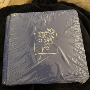 May include: A square, navy blue book wrapped in clear plastic. The cover features a white floral design within a white square. The book is on a black surface.
