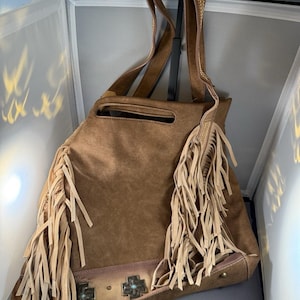 May include: A brown suede tote bag with long fringe and decorative metal cross accents. The bag has a shoulder strap with gold embroidery and a top handle. The bag has a large opening and a zippered pocket.
