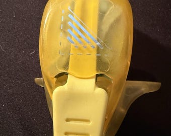 May include: A yellow craft punch with a clear plastic body and a light yellow handle. The punch has a design of blue lines within a dashed square. The handle has three horizontal grooves.