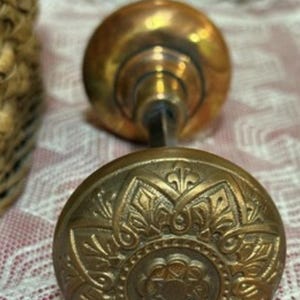 May include: Antique brass door knob set. The set includes two ornate, round knobs connected by a spindle. The knobs have a detailed, embossed floral design. The brass has a warm, aged patina, suggesting vintage charm.