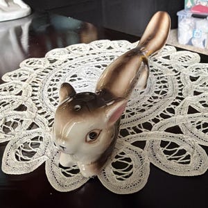 May include: A ceramic squirrel figurine featuring a glossy finish with shades of brown, tan, and white. The squirrel is depicted in a resting position with its tail extended upward. It has small, pointed ears with pink inner details and dark eyes.